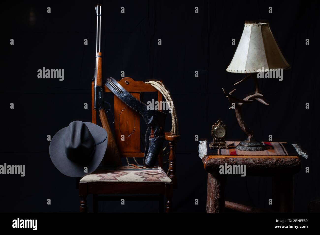 Lever action rifle, revolver, rope, cowboy hat, clock, table, lamp and a chair with a black background, horizontal Stock Photo