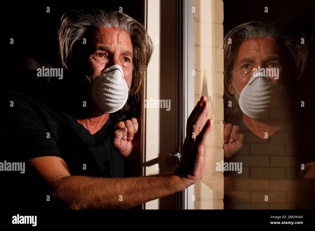 coronavirus crying male medical mask quarantine, self isolation concept, depressed distraught mans reflection in window with tears wearing protective Stock Photo