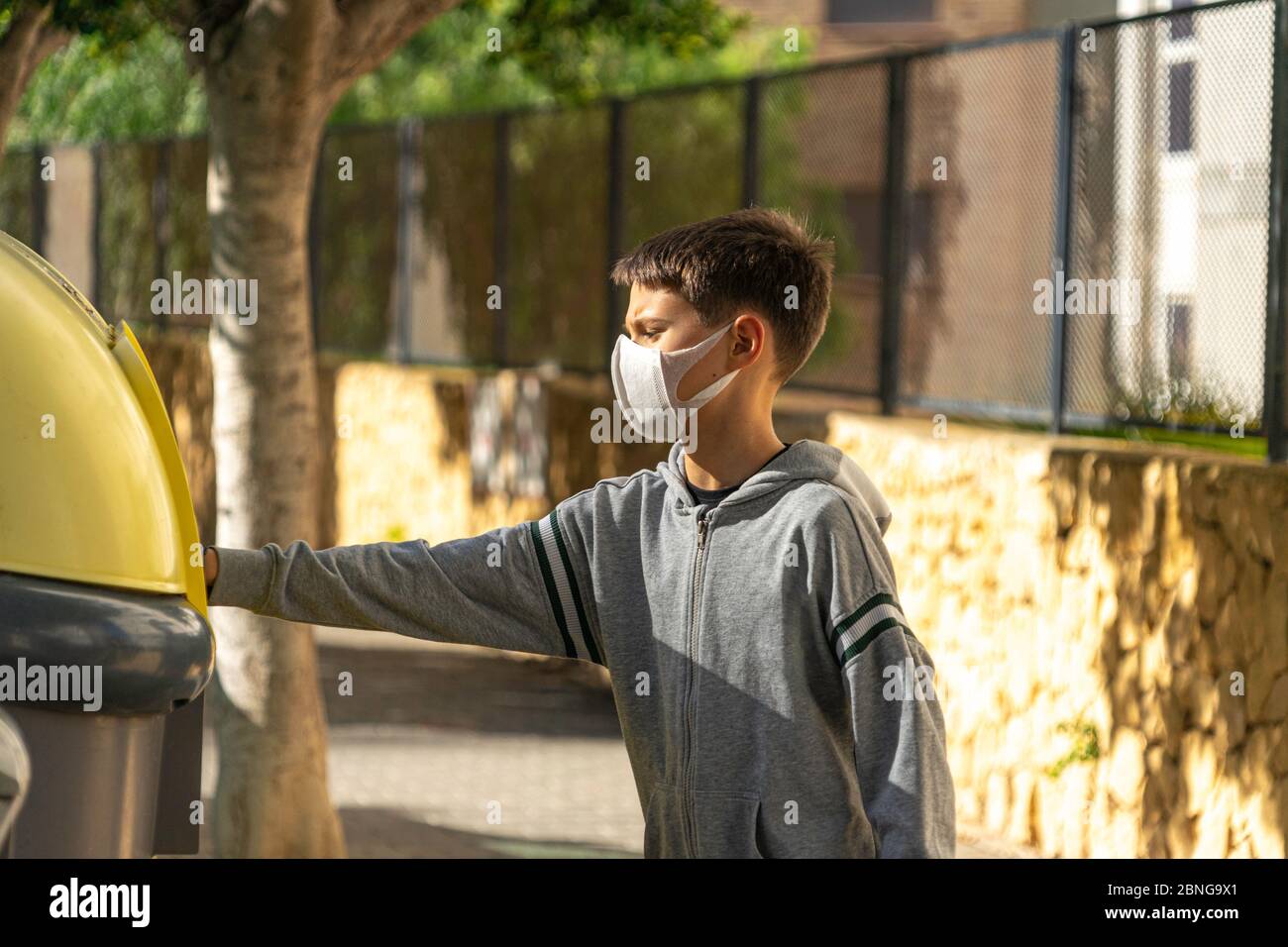 Child with protective mask and gloves throwing empty plastic bottle into recycling bin near residential apartment building outdoors Stock Photo
