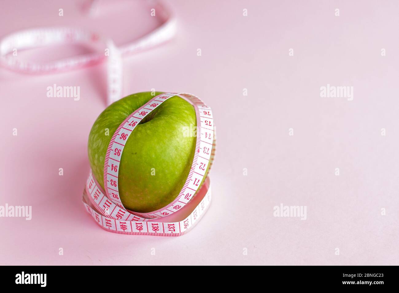 selective focus, green ripe Apple wrapped in measuring tape, the concept of proper nutrition and weight loss. on a pink background Stock Photo