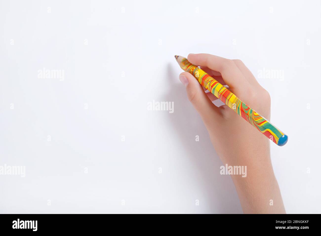 The child's hand holding a multicolored pencil against a white sheet of paper Stock Photo