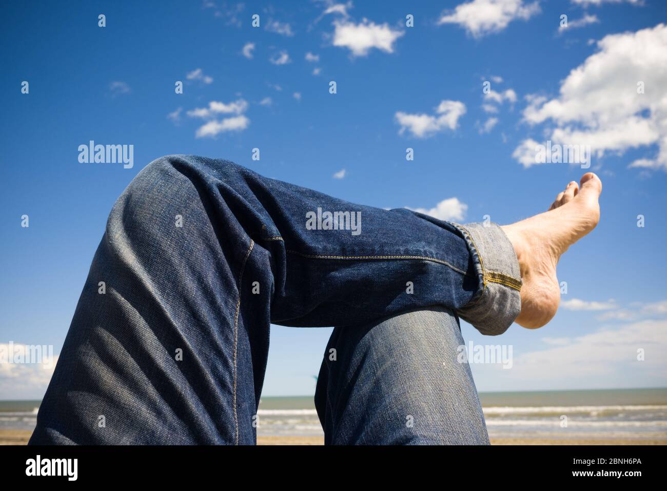 A Cropped View Of A Relaxing Man S Crossed Legs With Turned Up Blue Jeans And Bare Feet On A Sunny Beach With Sky Sandy Beach And Sea In Background Stock Photo