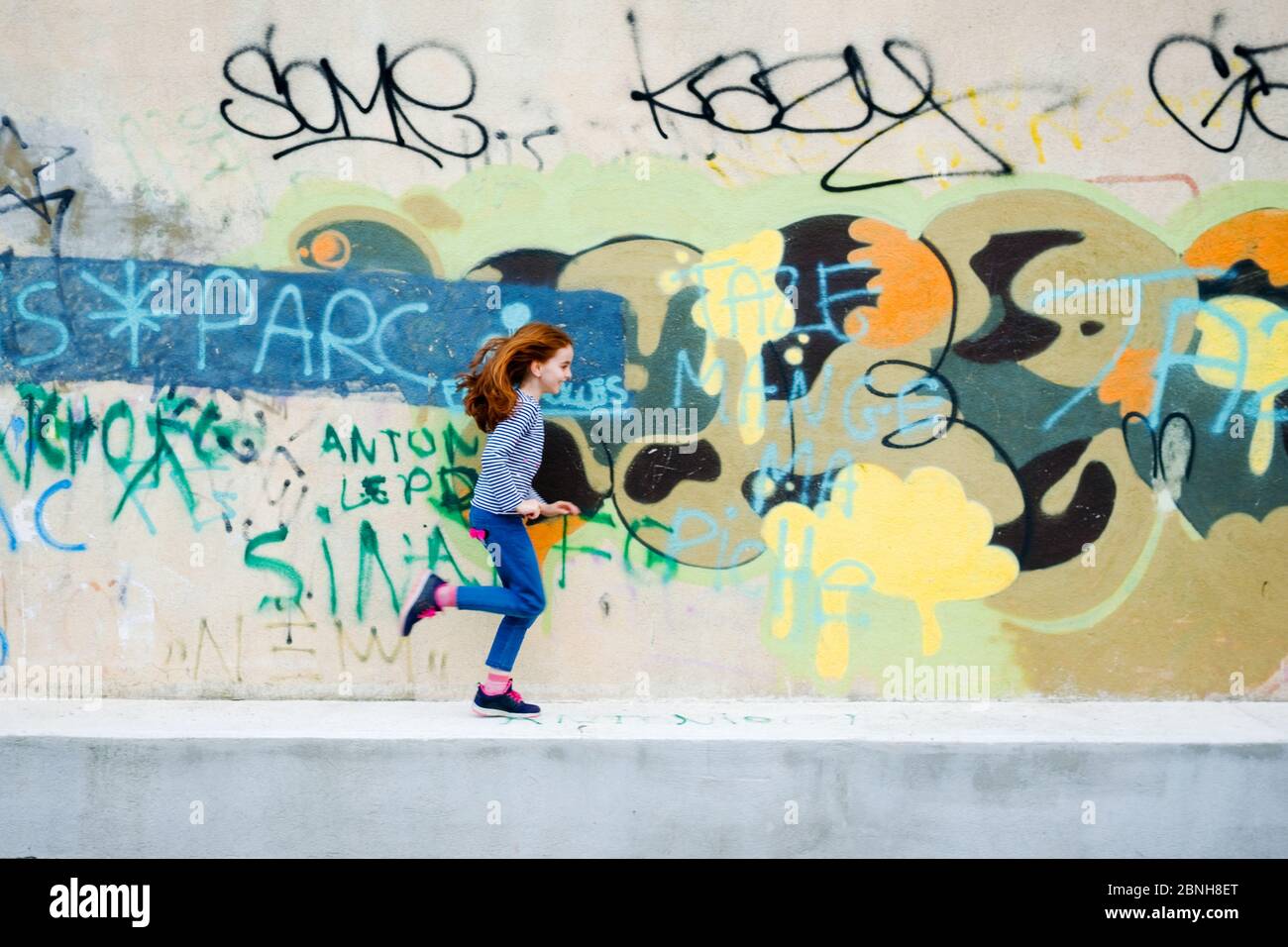 a seven year old girl with long red hair runs laughing in front of a wall covered with urban graffiti in Figuerolles, Montpellier, France Stock Photo