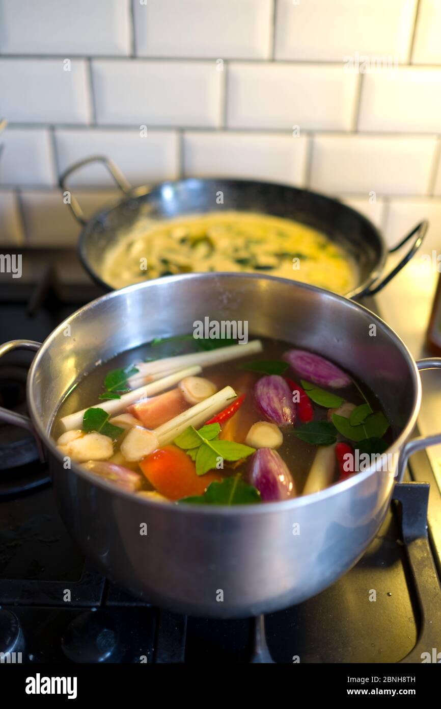 a close up of a cooking pot on the hob with a thai soup with fresh vegetables and a dish with curry in the background in a domestic kitchen Stock Photo
