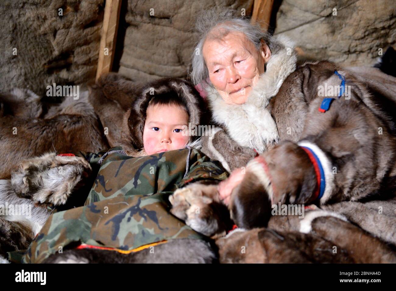 Nenet elder with grandson dressed in winter coats, made from reindeer skin in tent. Yar-Sale district, Yamal, Northwest Siberia, Russia. April 2016. Stock Photo