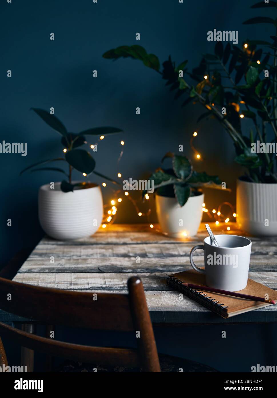Cozy workspace with note book with and cup of coffee at wooden table with plants and yellow glowing garland at stone grey wall background. Working fro Stock Photo