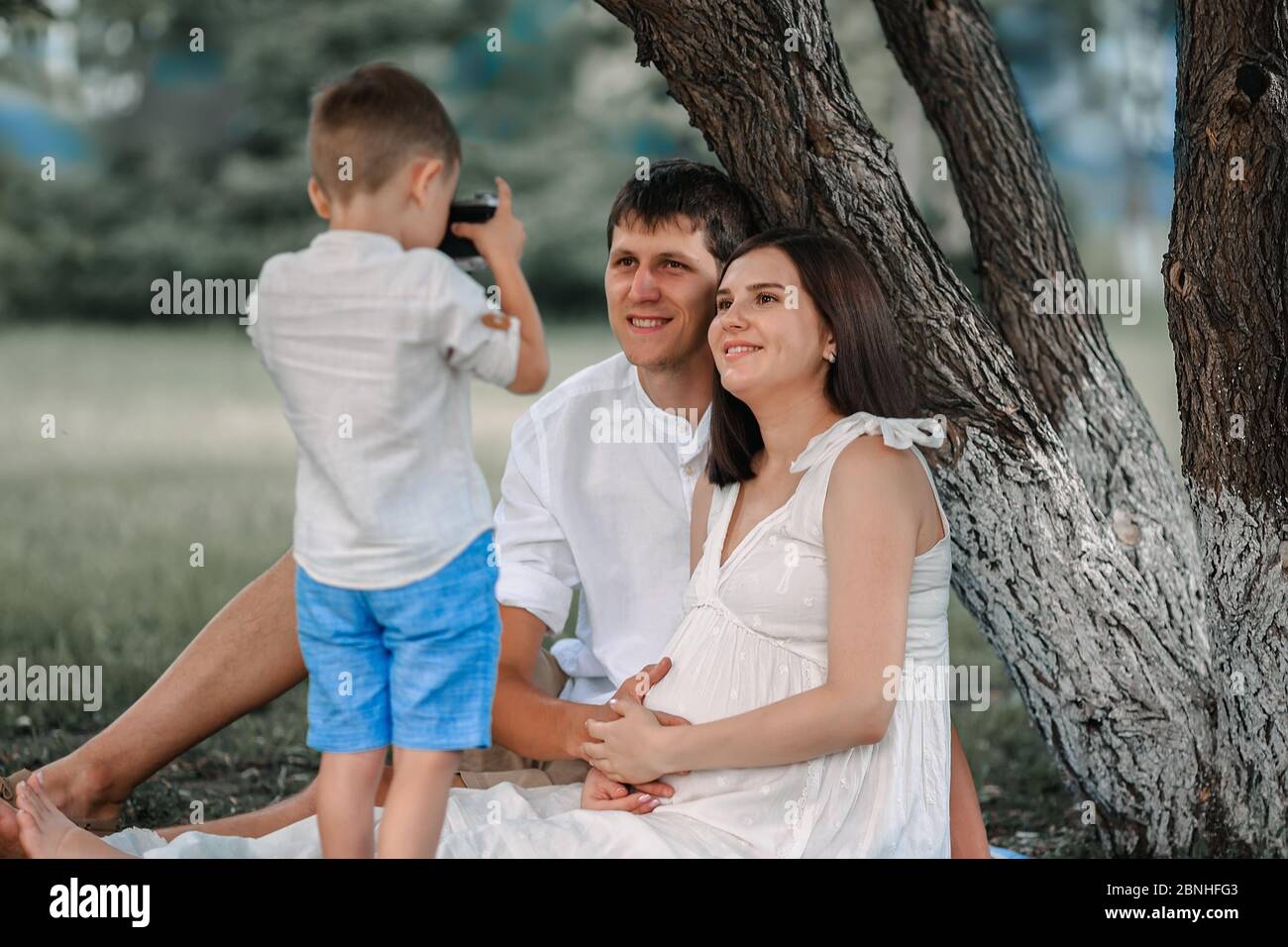 Happy family relaxes in nature under a tree in the afternoon. The child takes pictures of his father and pregnant mother Stock Photo