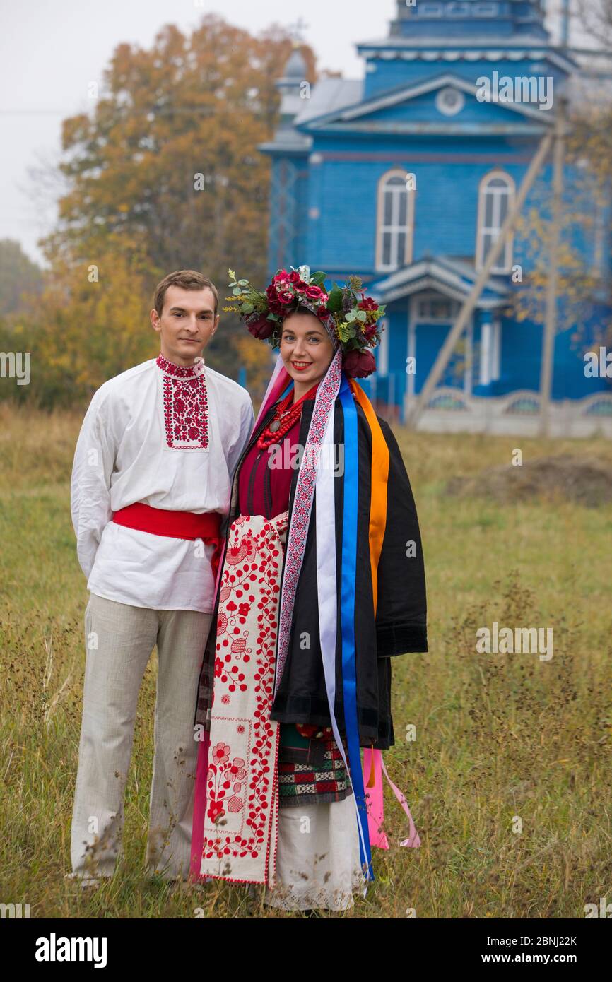 Girl and guy in retro costumes on the street in the old village. Retro staging of an ancient rite. Beautiful wreath on a girl. The guy hugs the girl Stock Photo