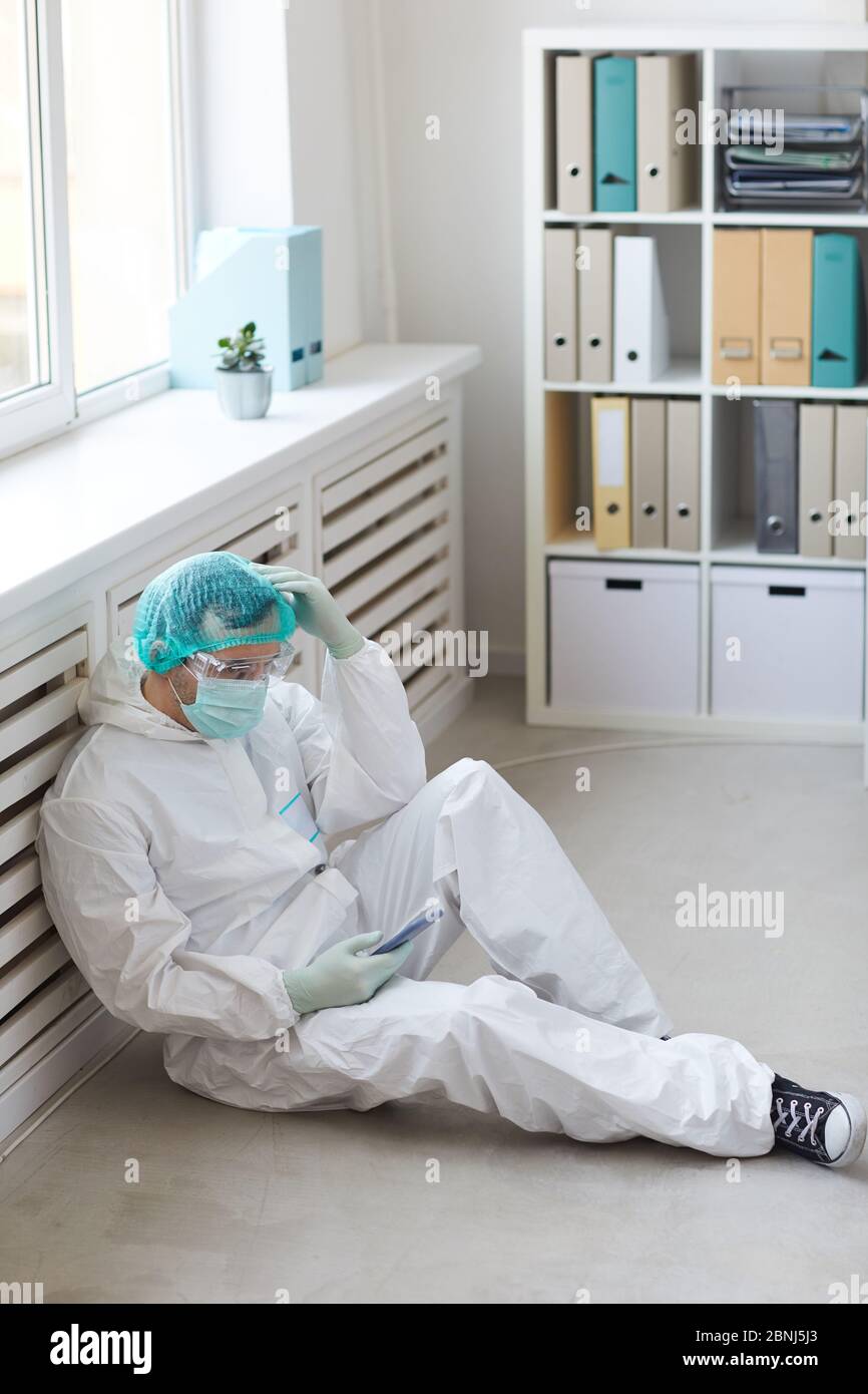 Male doctor in protective clothing sitting on the floor and using his mobile phone he is tired and resting Stock Photo