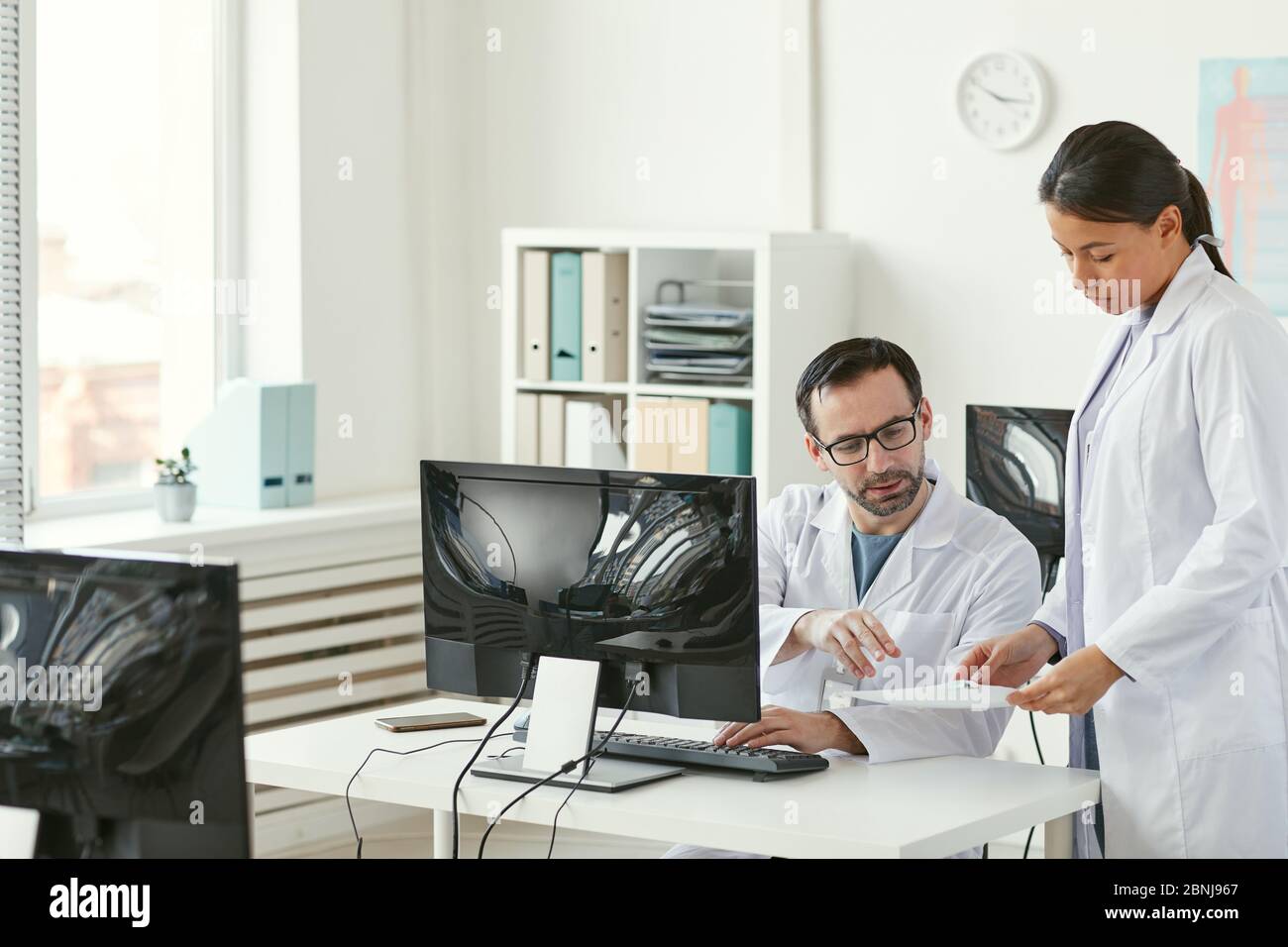 Mature male doctor sitting at his workplace in front of computer and discussing documents with nurse at office Stock Photo