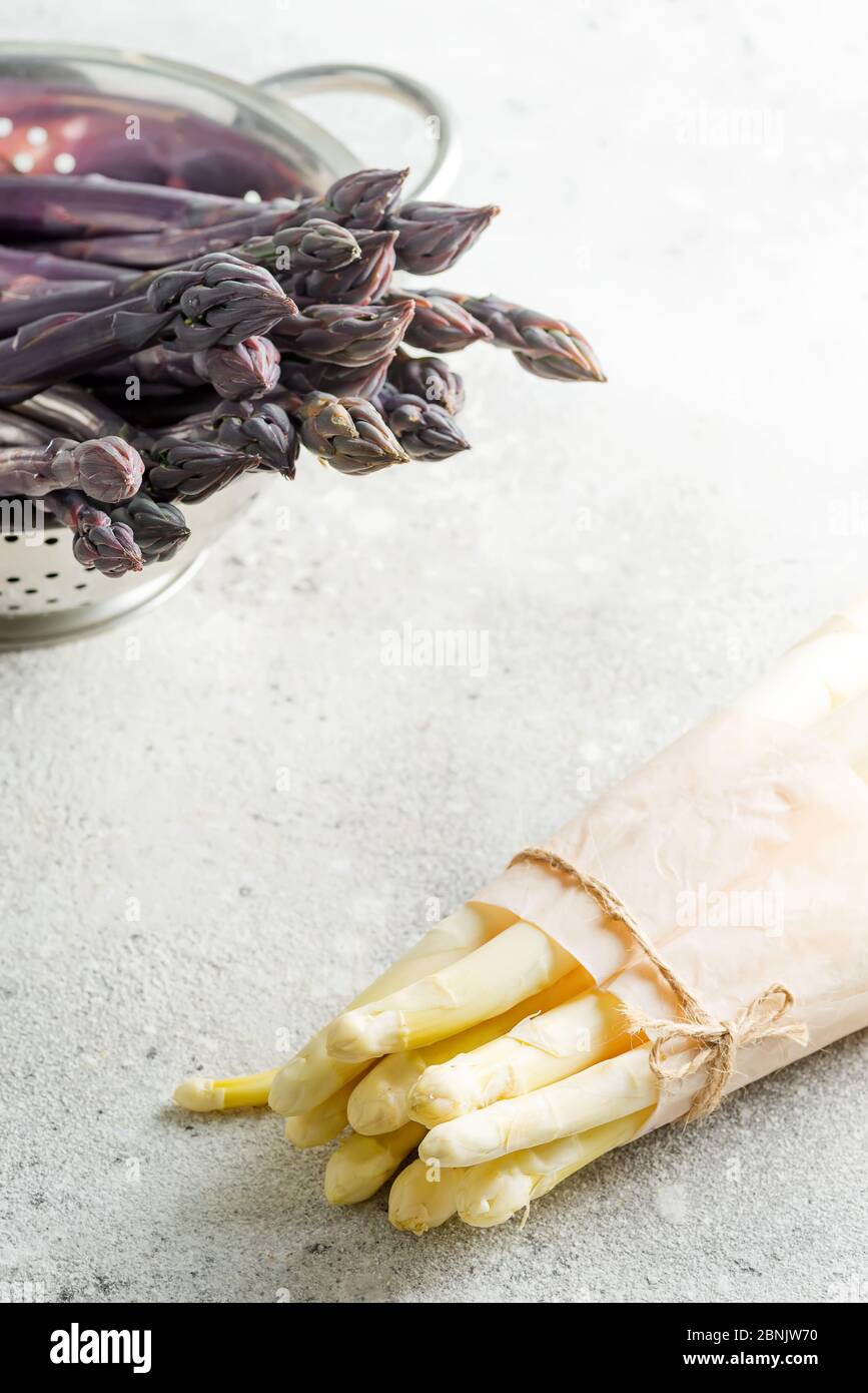 Raw organic purple asparagus spears in colander and bunch of white asparagus as a healthy diet food on a light stone background. Stock Photo