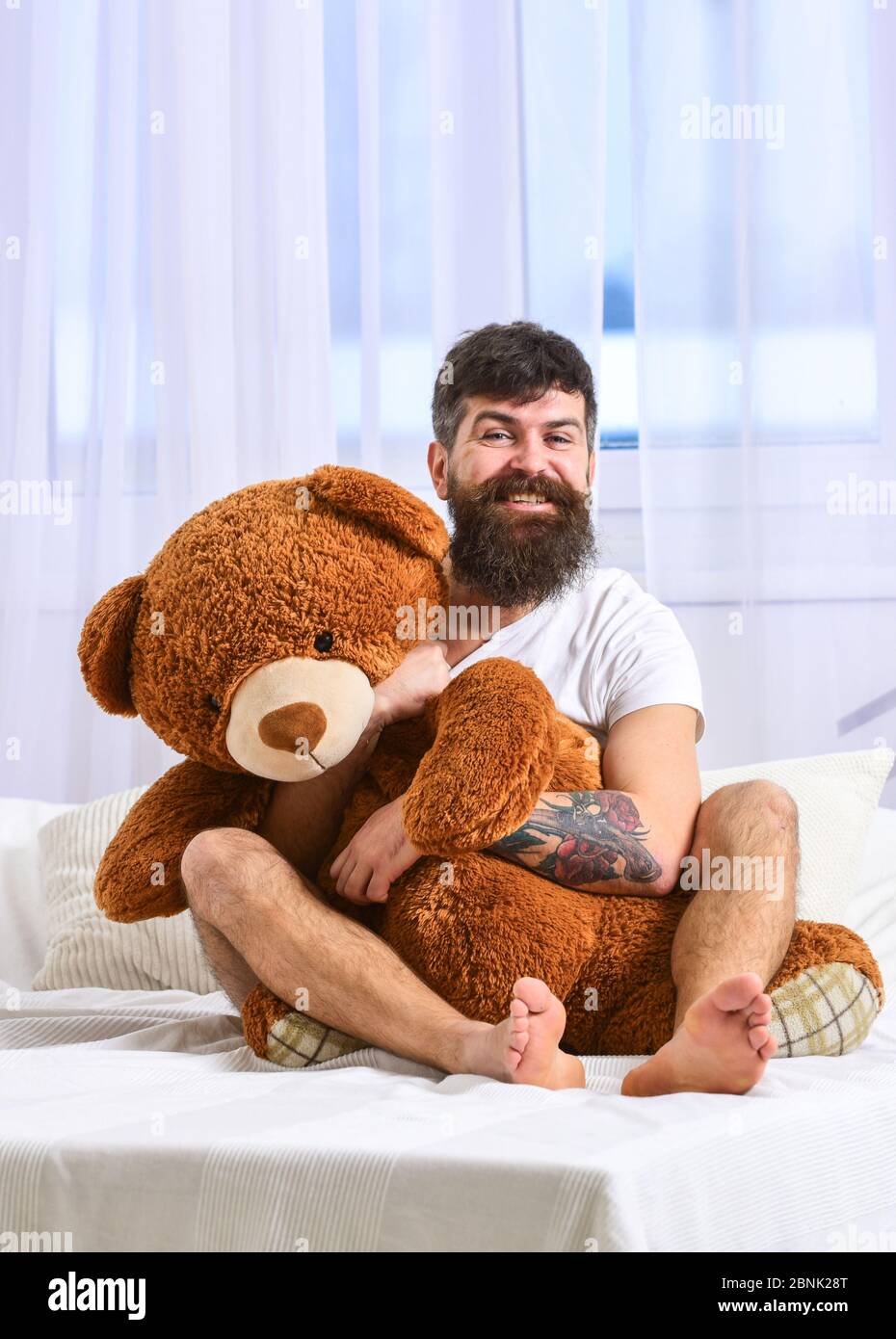 Guy on cheerful face hugs giant teddy bear. Birthday concept. Macho with beard and mustache cuddling with plush toy on birthday morning. Man sits on bed and hugs big toy white curtains