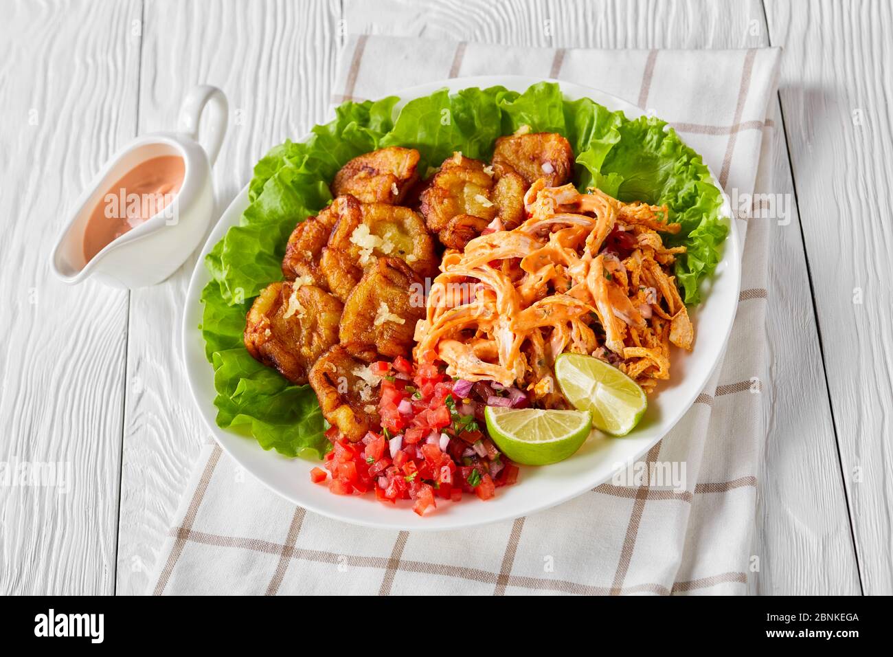 Easy spicy fried plantains served with shredded chicken mixed with buffalo sauce, pico de gallo salsa, lime wedges on fresh lettuce leaves, on a white Stock Photo