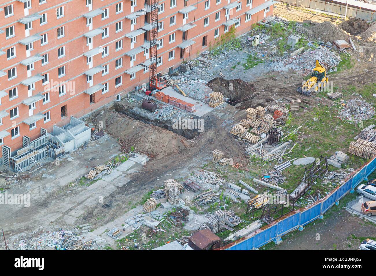 Fenced territory with a concrete blue fence for the construction of a red brick apartment building with a crane and an excavator with dug ground for c Stock Photo