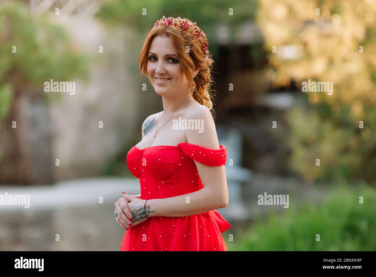 Portrait of attractive smiling redhead tattooed woman in red dress and diadema on her head posing on blurred medieval castle background at summer suns Stock Photo