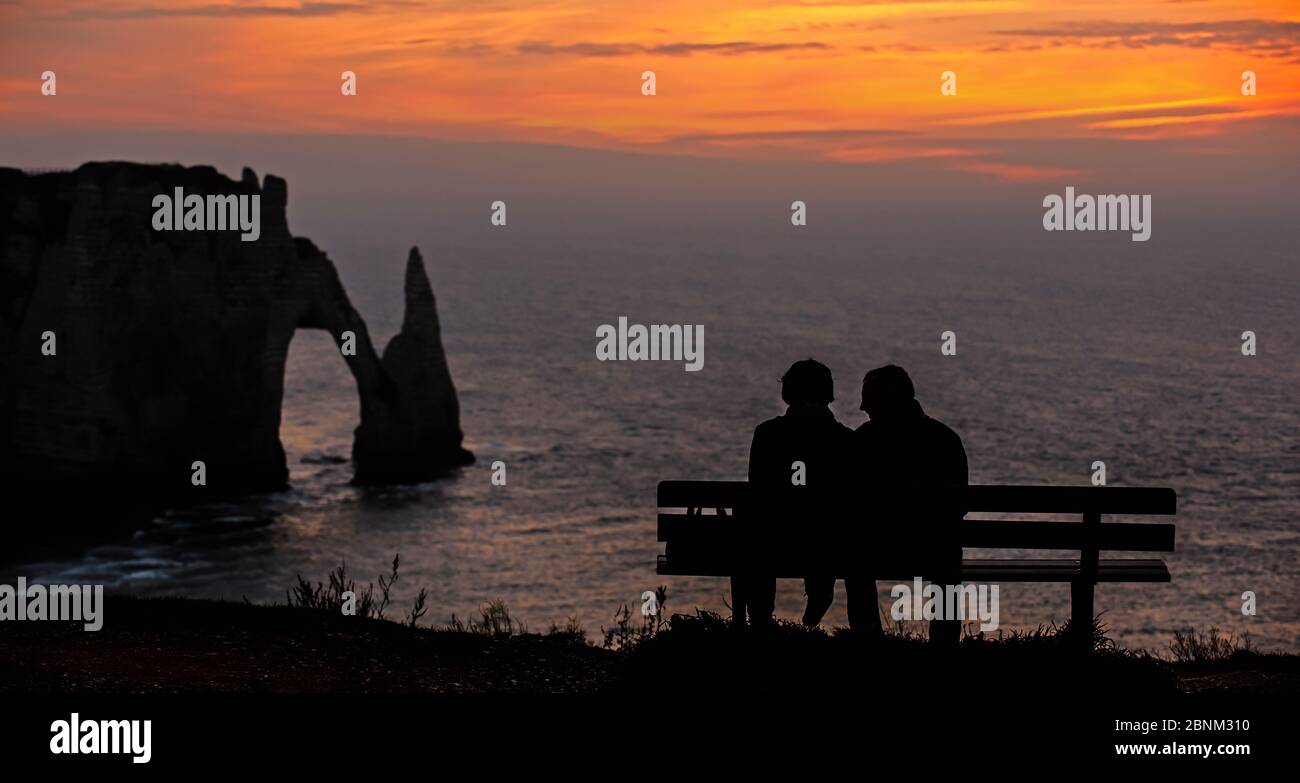 Elderly tourists sitting on bench looking at the Porte D'Aval, a natural arch in the chalk cliffs at Etretat at sunset, Cote d'Albatre, Upper Normandy Stock Photo