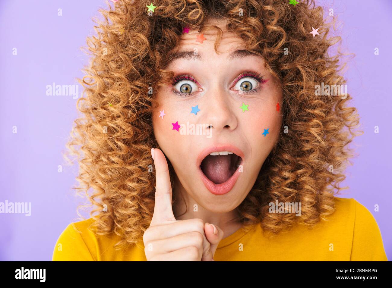 Image of pretty curly girl having idea and pointing finger upward with multicolored glitter