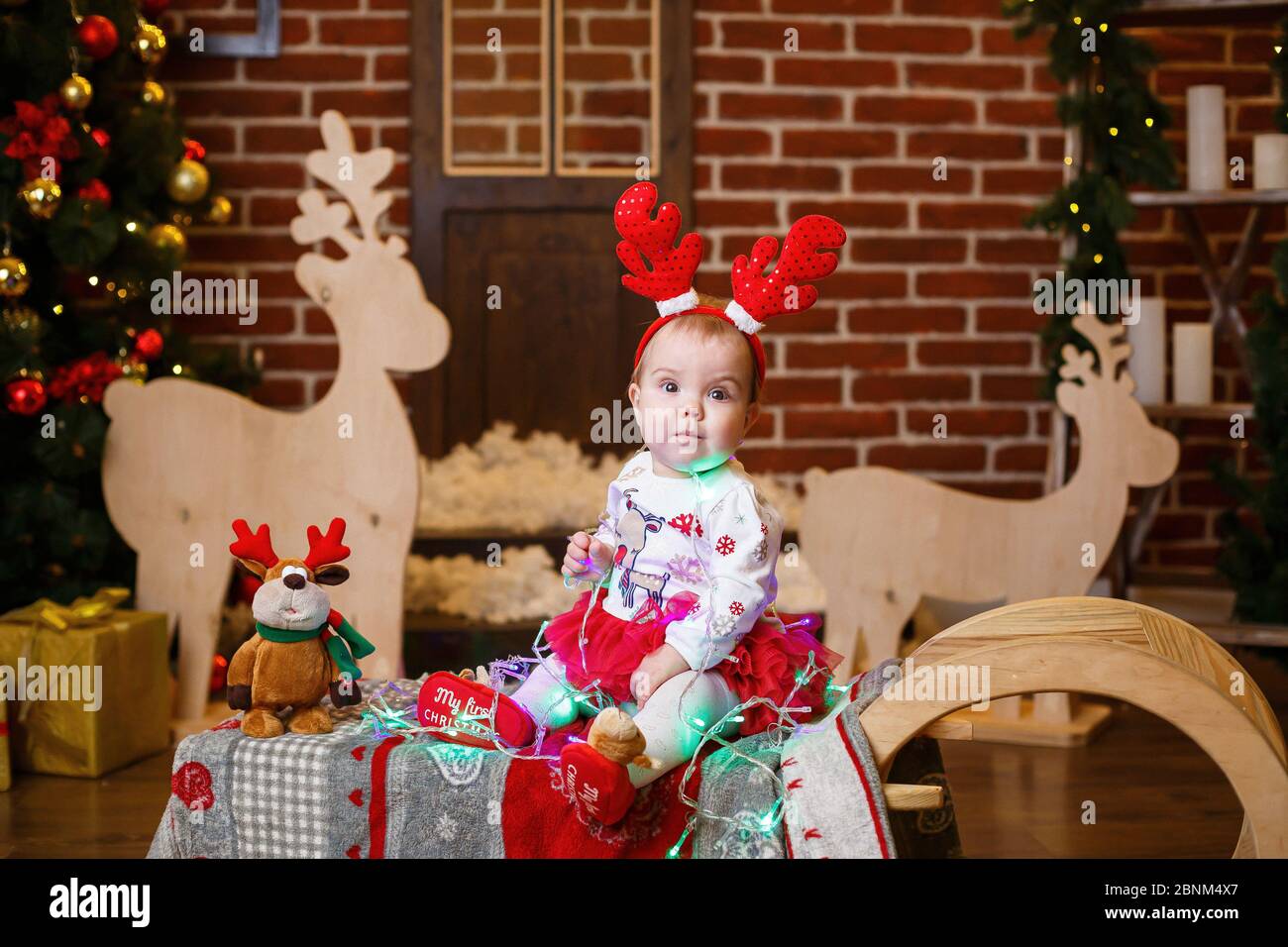 A little girl in warm sweater sits under a Christmas tree with toys and gifts with horns on her head. Happy childhood. New Year holiday atmosphere Stock Photo