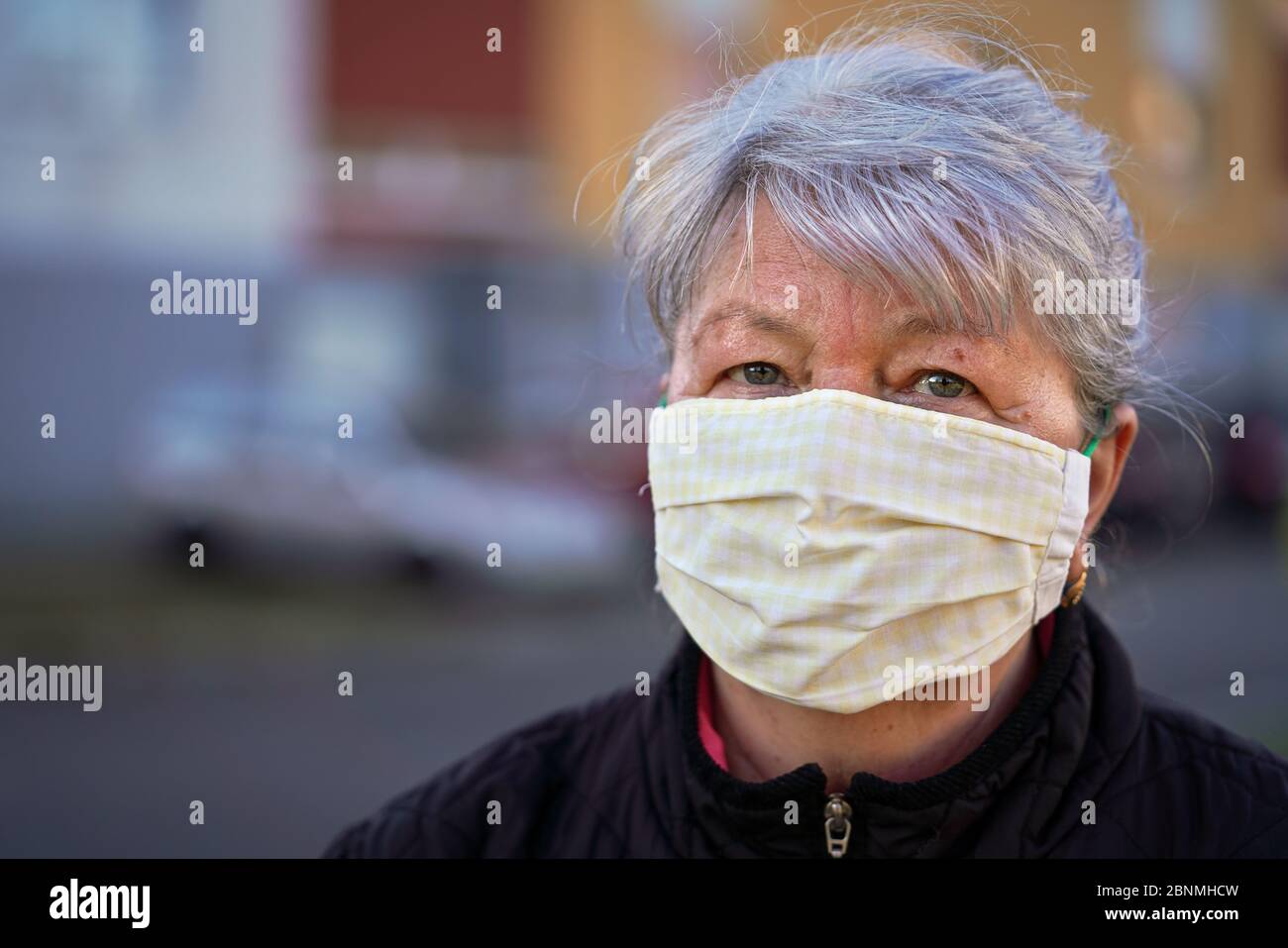 Elderly senior woman wearing home made cloth face mouth nose virus mask outside, blurred building background. Can be used during coronavirus covid-19 Stock Photo