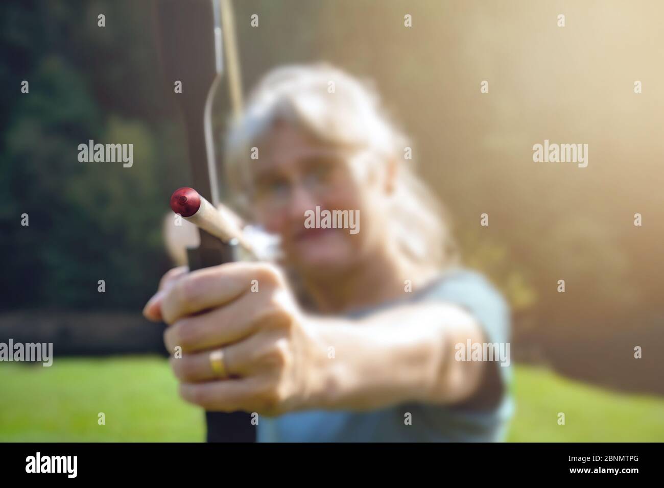 Woman with gray hair aiming with bow and arrow in nature and sun. Concept for the fight to reach the target Stock Photo