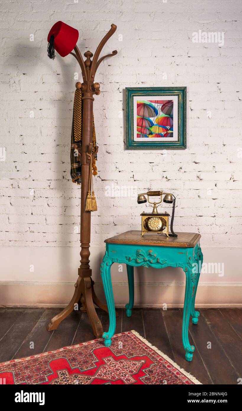 Green wooden vintage side table with golden antique telephone set, and coat hanger stand with red fez and scarf, on room with white bricks wall, hanged painting, and wooden parquet floor Stock Photo