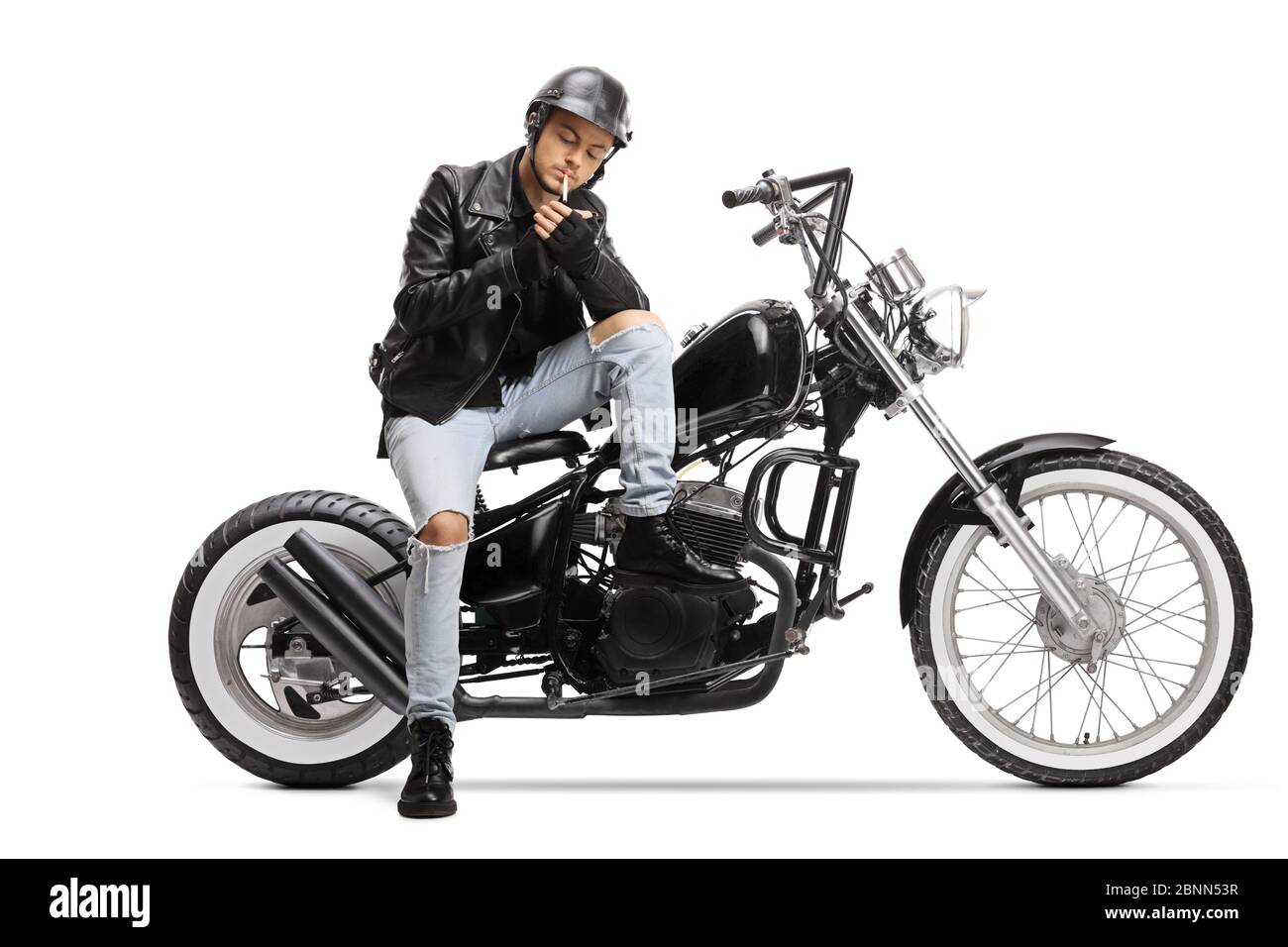 Young biker with a helmet and a leather jacket sitting on a chopper motorbike and lighting a cigarette isolated on white background Stock Photo