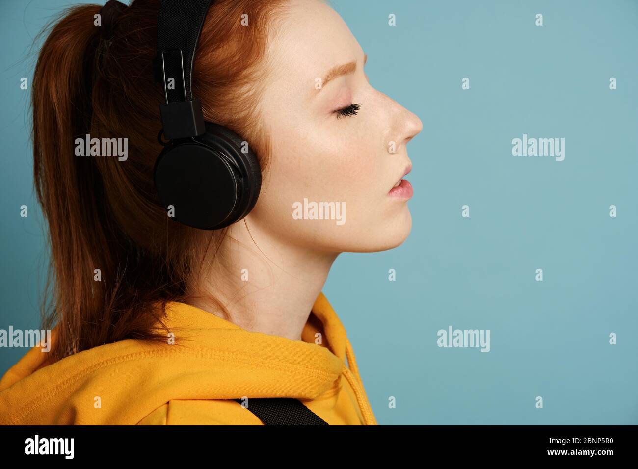 Head shot. A red-haired girl wearing headphones and a yellow hoodie stands on a blue background in a profile with her eyes closed Stock Photo
