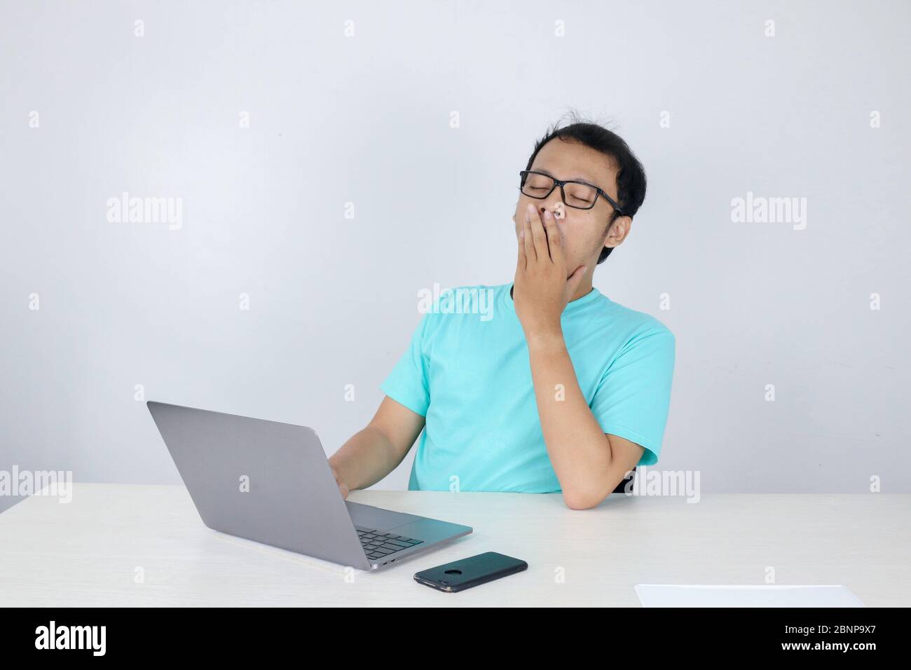 Young Asian Man is Lazy and tired with sleep when working in the front of Laptop. Indonesian man wearing blue shirt. Stock Photo
