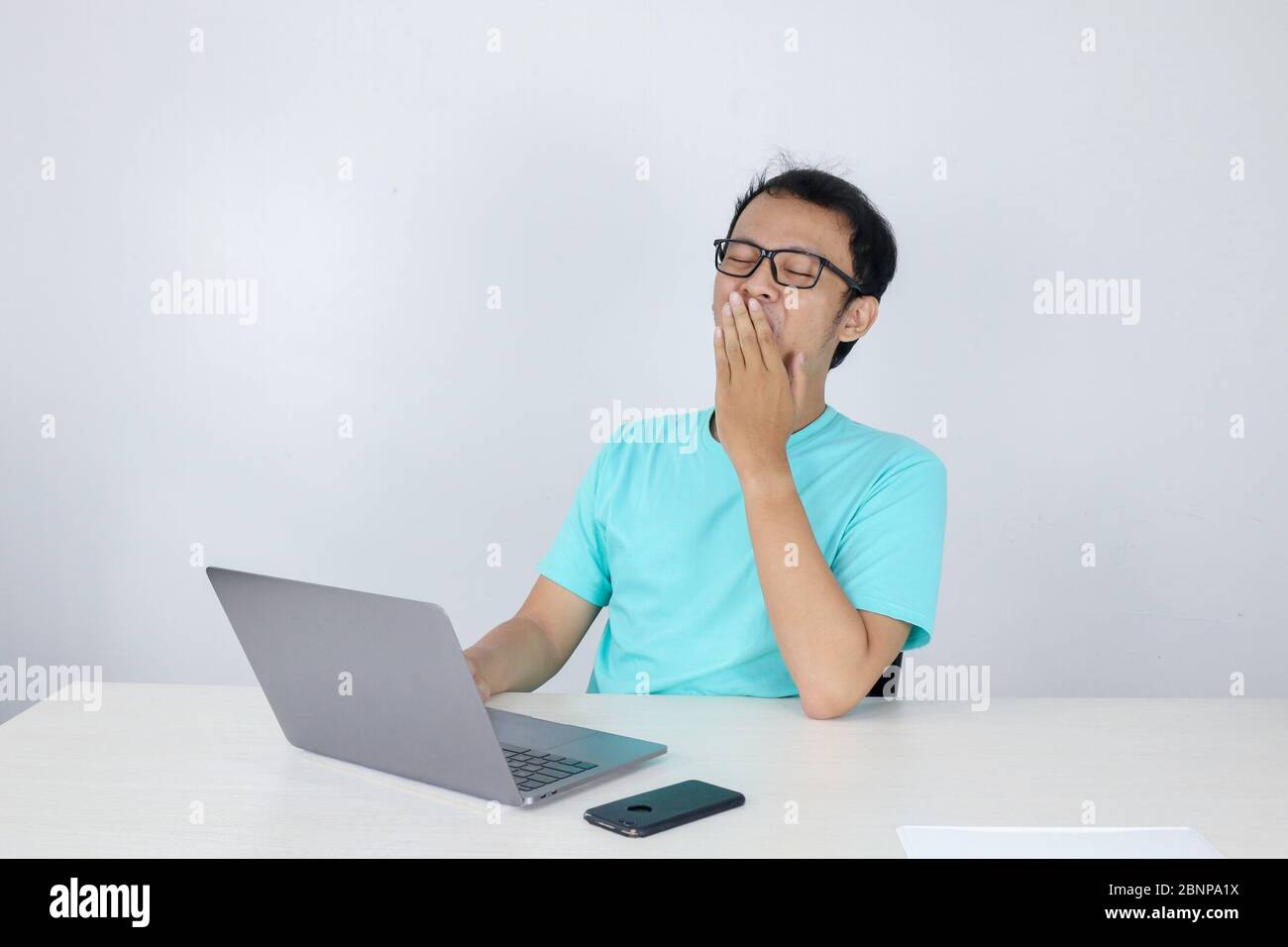 Young Asian Man is Lazy and tired with sleep when working in the front of Laptop. Indonesian man wearing blue shirt. Stock Photo