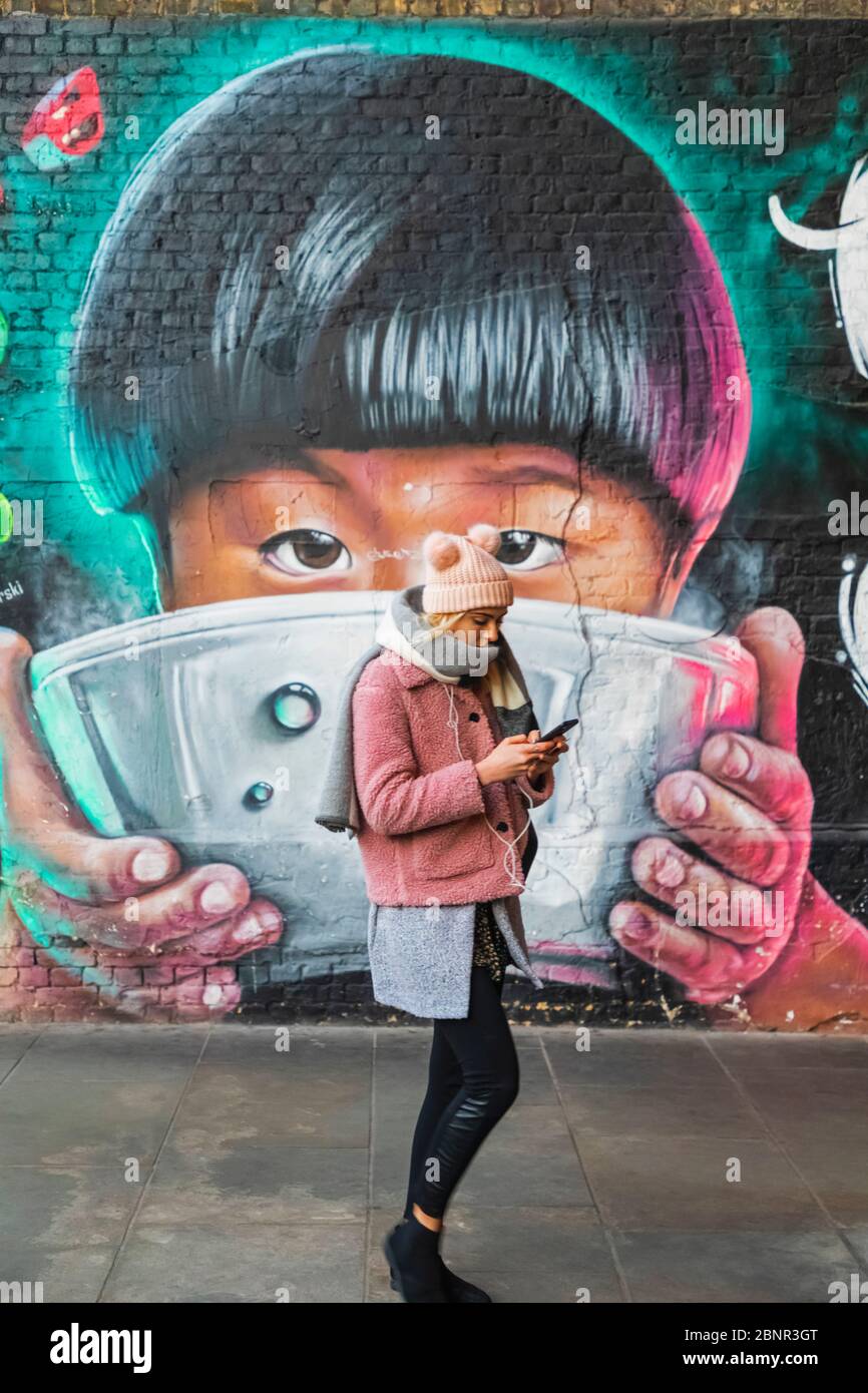 England, London, Southwark, Clink Street, Girl Texting While Walking Past Wall Mural Street Art depicting Asian Child Eating from Bowl Stock Photo