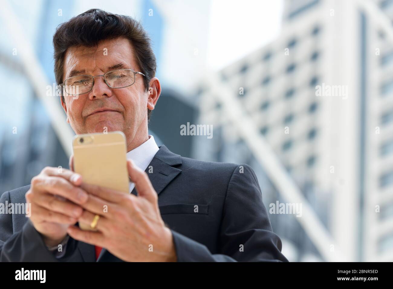 Mature businessman using mobile phone in front of modern building at ...