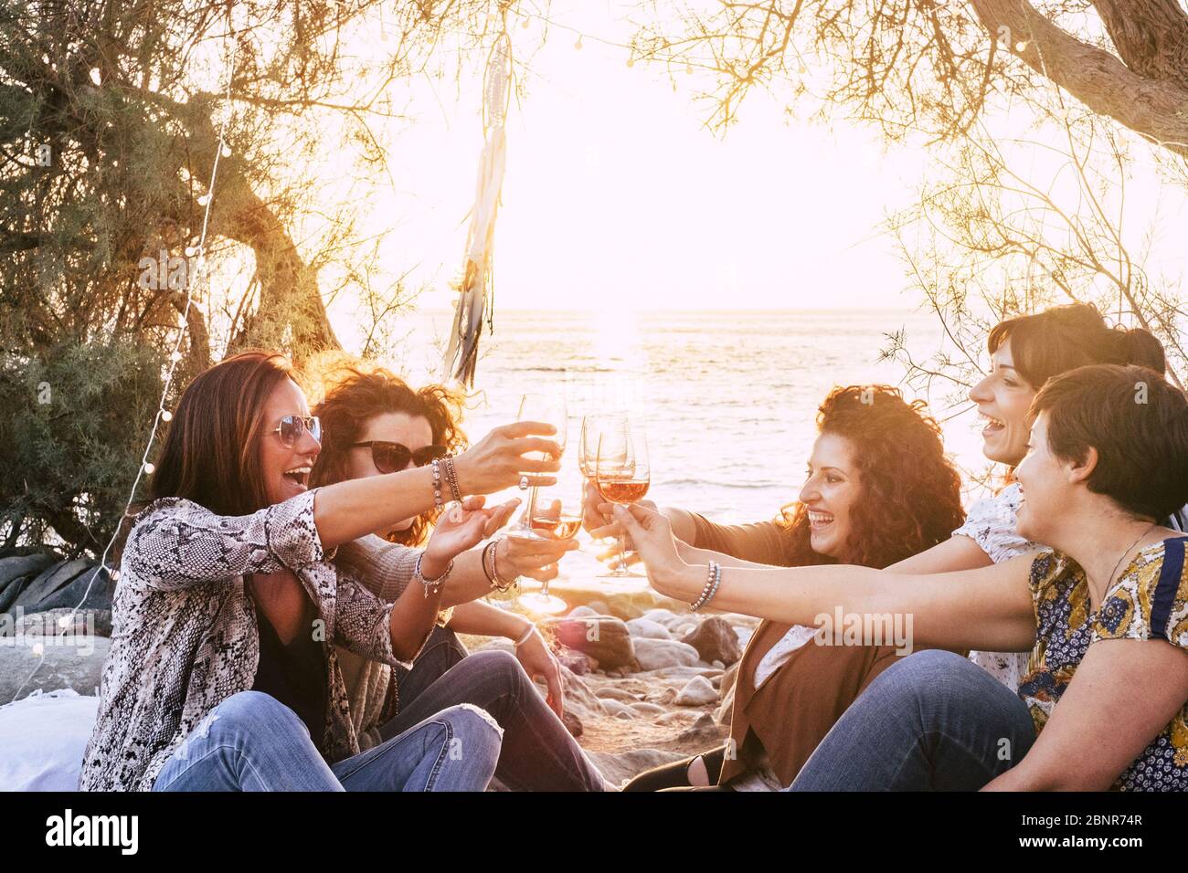 Group of middle age young adult women having fun together toasting and clinking with wine glasses at the beach during a golden sunset enjoying outdoor leisure activity or vacation Stock Photo