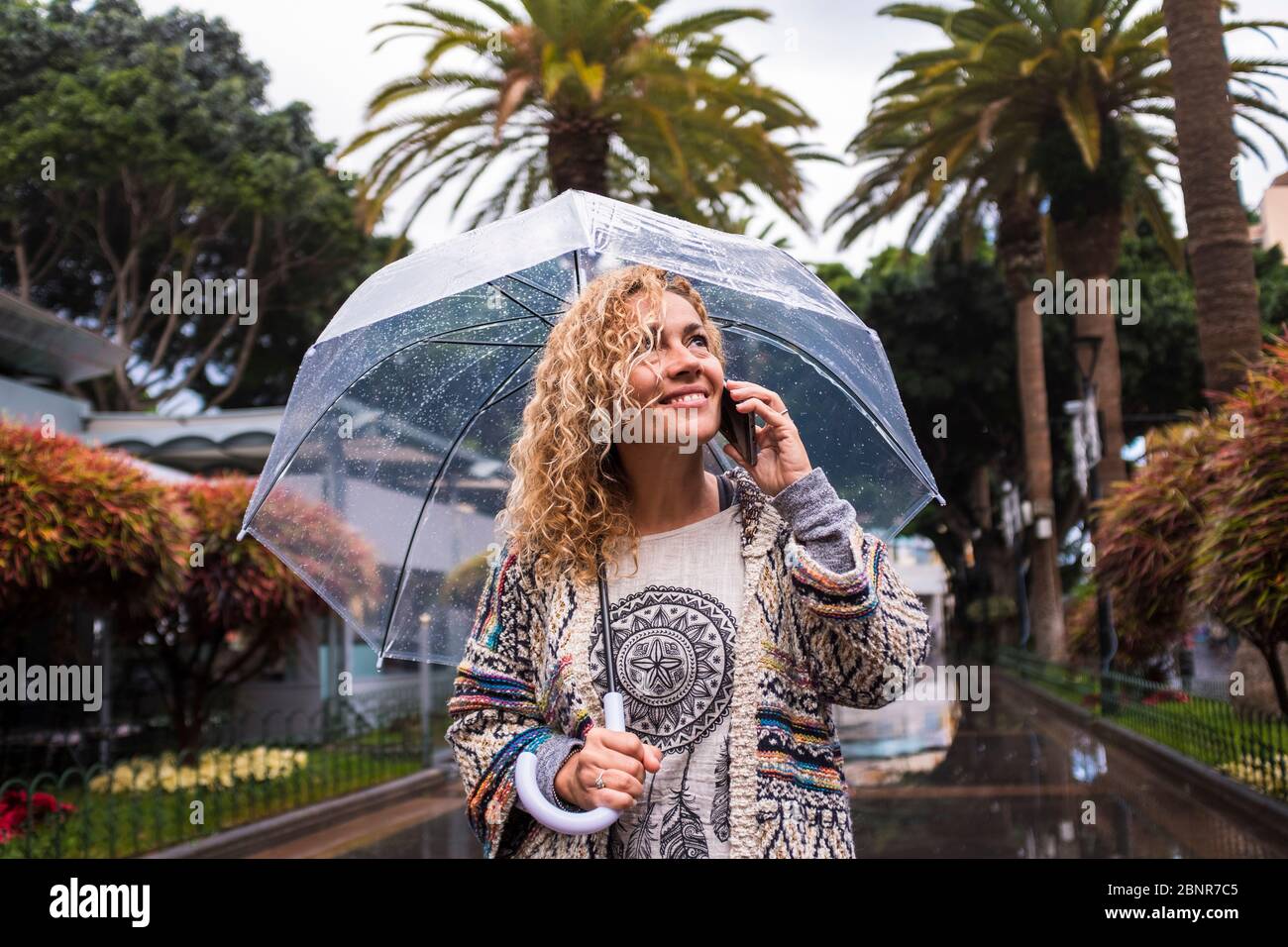 Attractive and cheerful fashion trendy middle age young caucasian woman walking in the city under the rain with a transparent umbrella - cold season concept enjoying the urban park Stock Photo