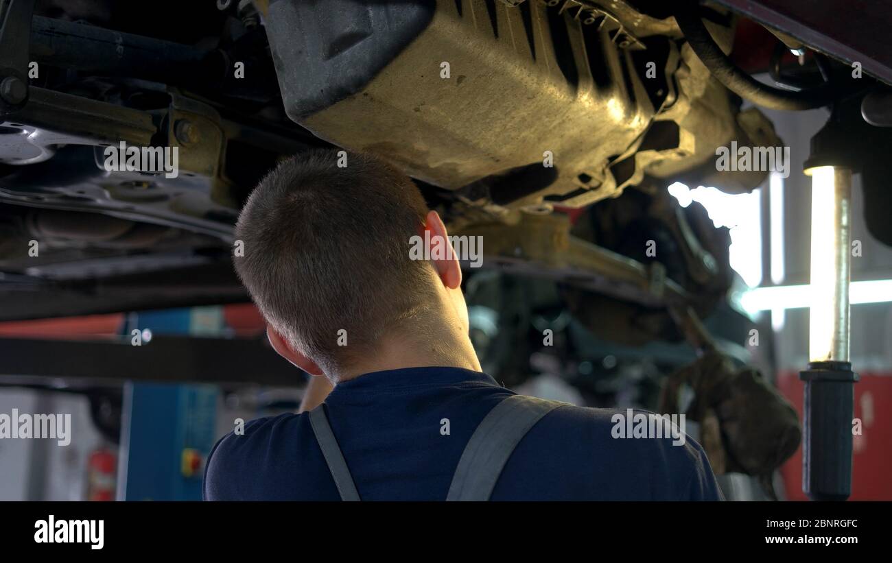 Young auto mechanic standing under lifted car bottom and unscrewing bolts. Stock Photo