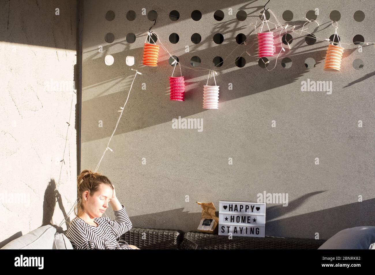 balcony decorated with colorful lanterns, woman sitting in the sun, sign 'happy home staying' Stock Photo