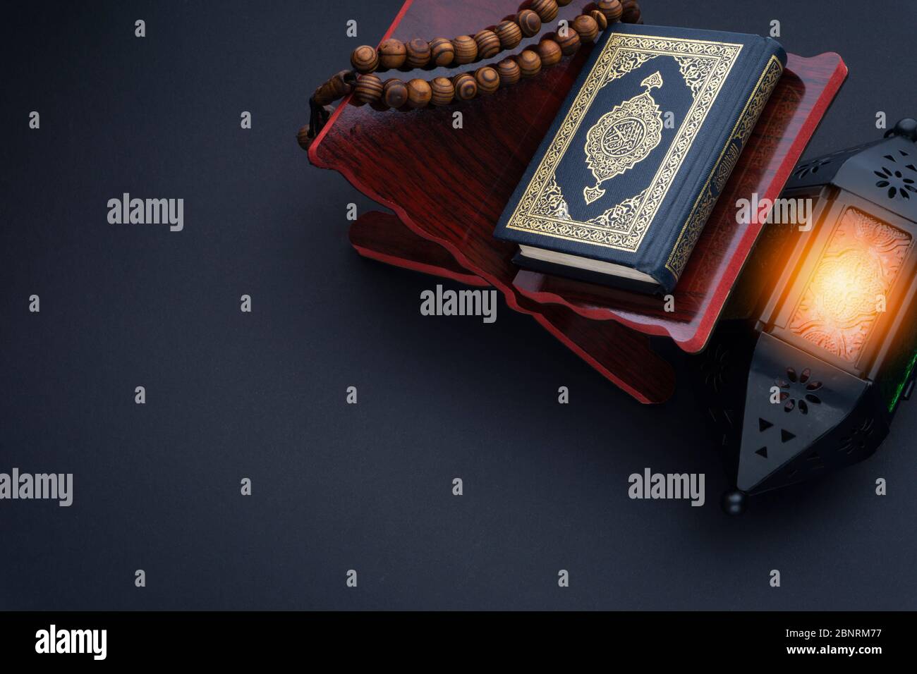Holy Al Quran with written arabic calligraphy meaning of Al Quran, lantern lamp and rosary beads or tasbih on black background. Stock Photo