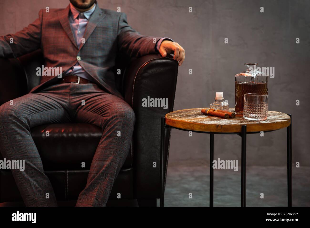 Gentleman sits in a leather armchair with carafe of whisky, empty glass and cigar on the table. Stock Photo