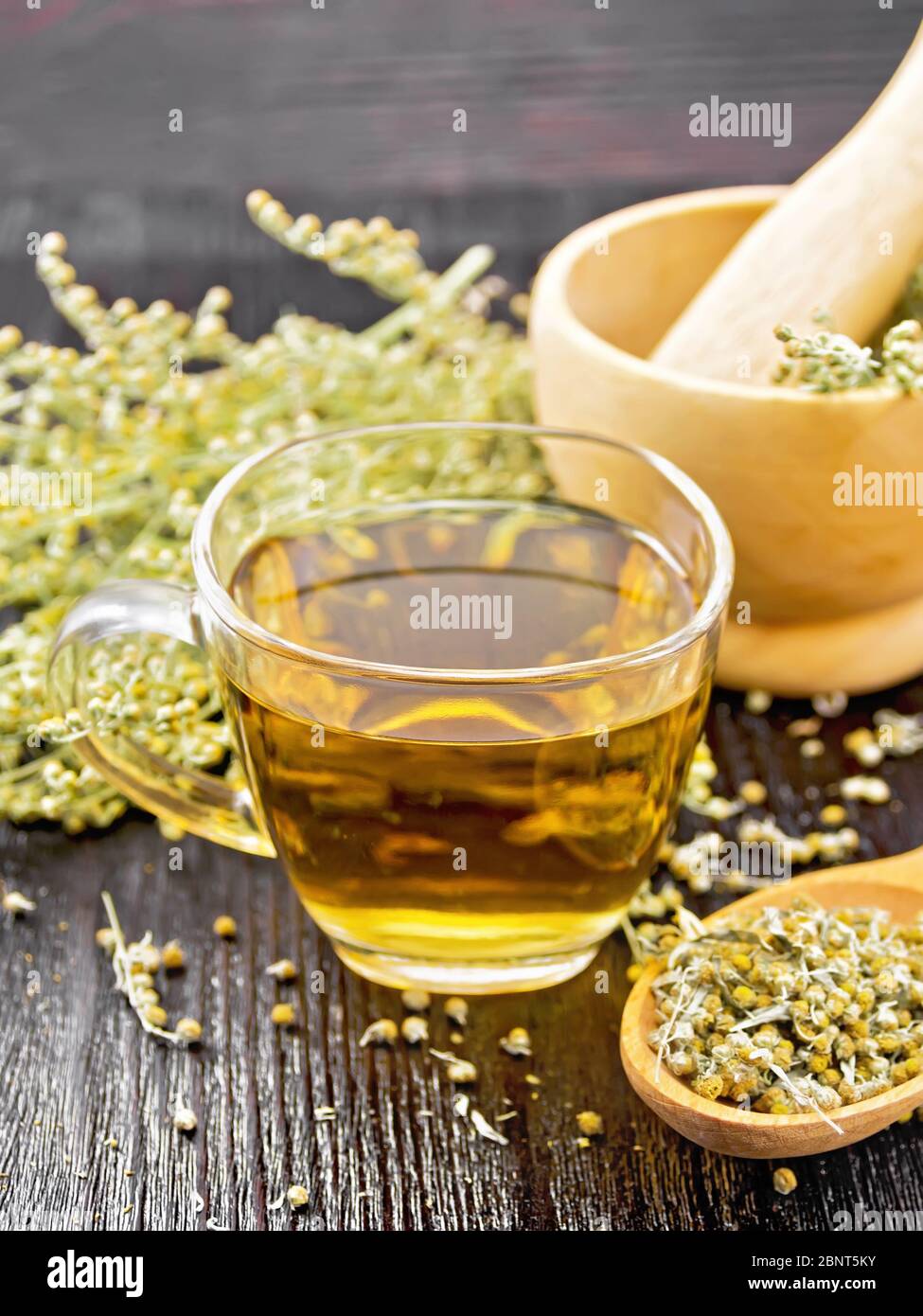 Gray wormwood herbal tea in a glass cup, dry sagebrush flowers in spoon, fresh flowers in mortar and on table on wooden board background Stock Photo