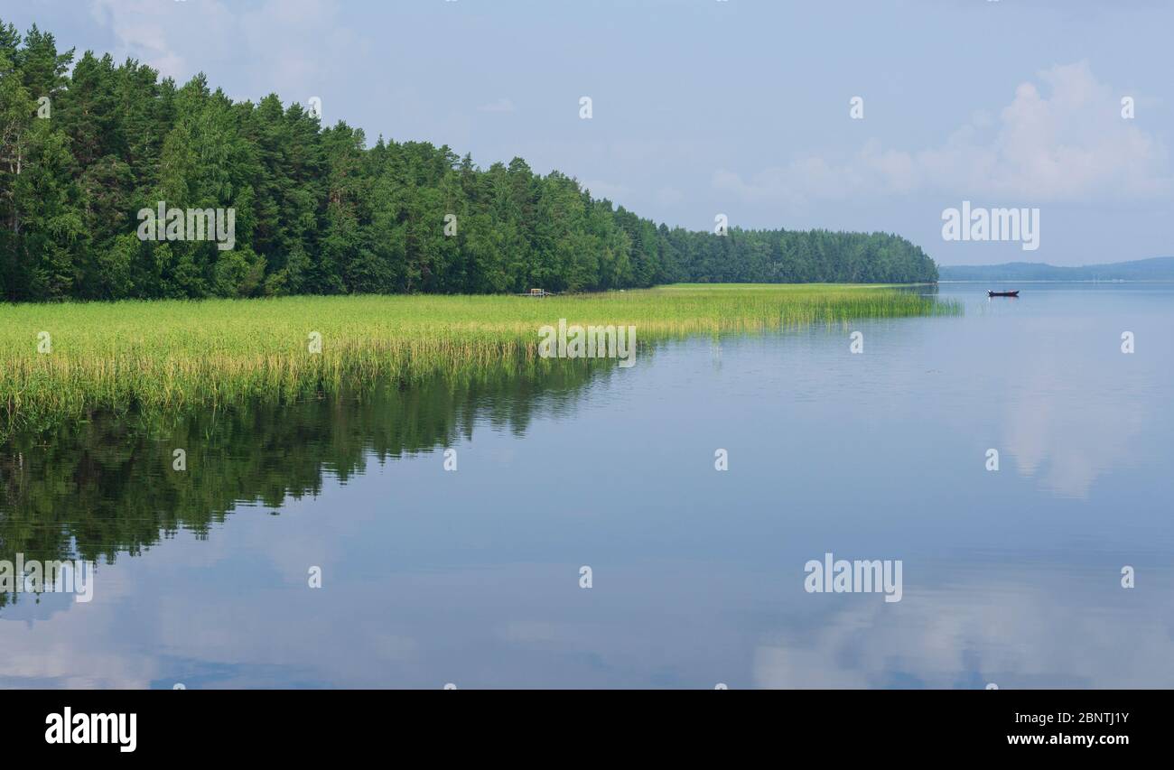 View of windless Lake Pohjois-Konnevesi, a cape growing trees and a distant rowboat / skiff at the lake , Finland Stock Photo