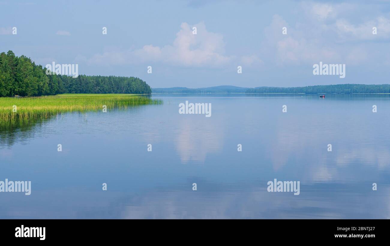 View of windless Lake Pohjois-Konnevesi, a cape growing trees and a distant rowboat / skiff at lake , Finland Stock Photo