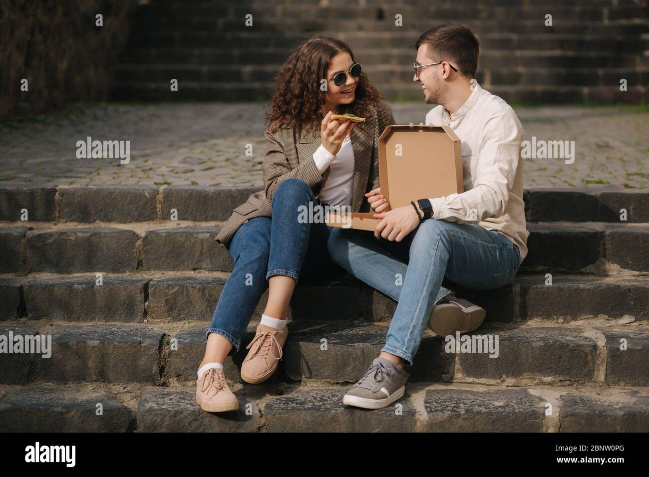 Young couple sitting on stairs outdoor and eating pizza. Handsome man with beaufitul woman taste pizza with vegetables. Vegan fast food Stock Photo