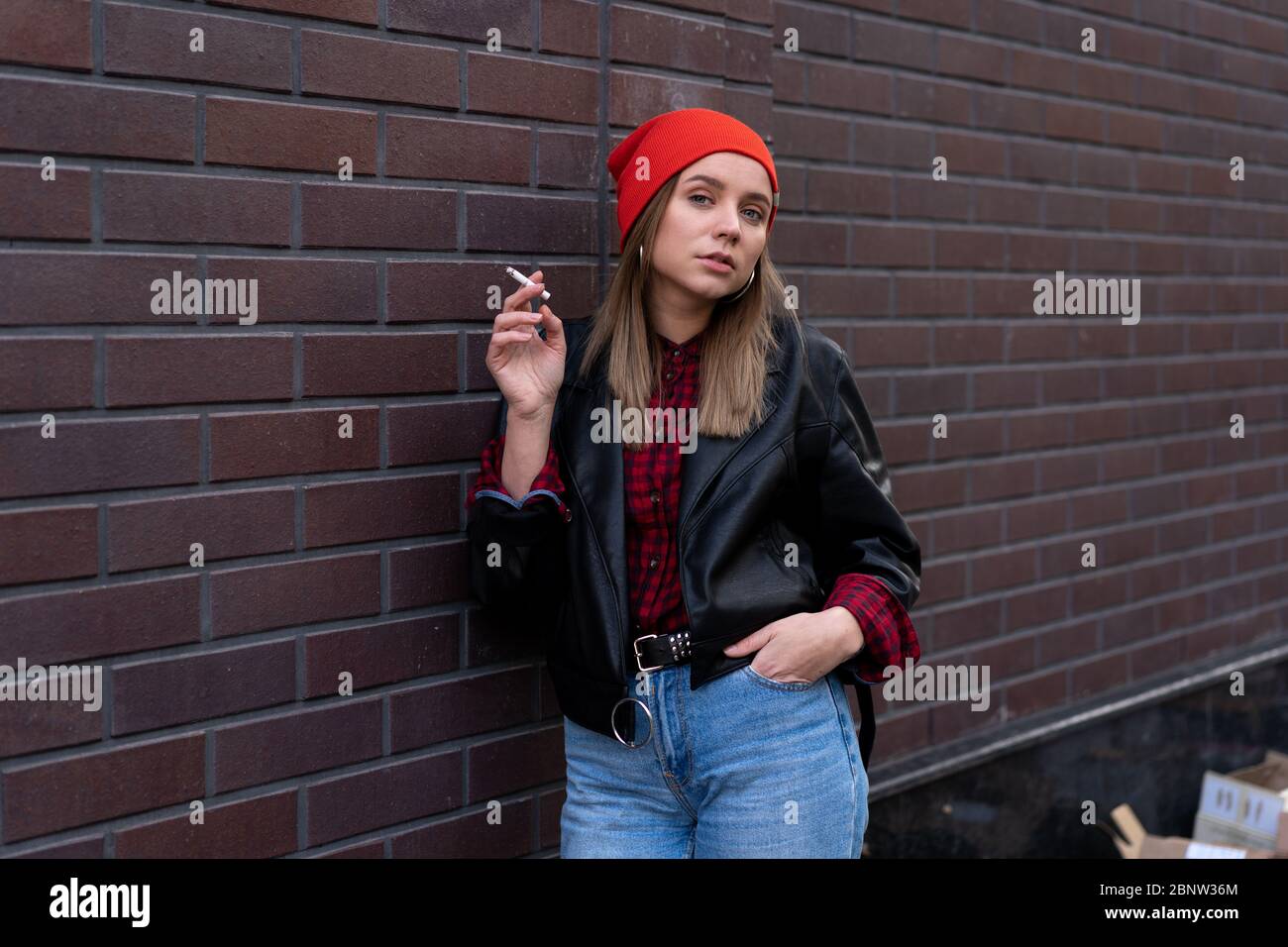 Beautiful young stylishly dressed Caucasian girl smokes cigarette on the street. Smoking bad habit Nicotine addiction Unhealthy lifestyle.  Modern wom Stock Photo