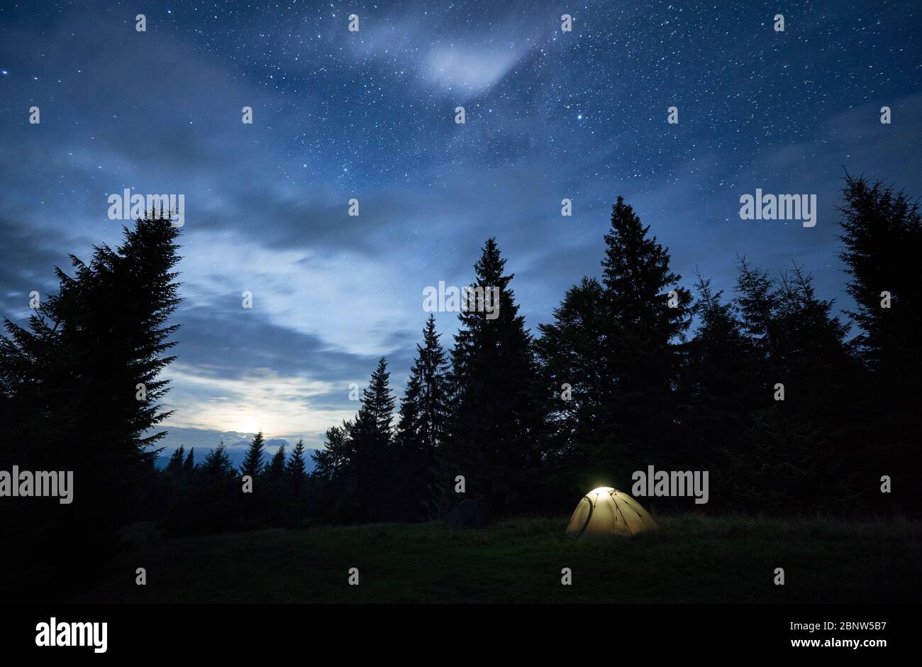 Fantastic View Of Night Starry Sky Over Meadow Stunning Scenery Of Tourist Illuminated Tent In Forest With Coniferous Trees Under Blue Sky With Stars Concept Of Travelling Hiking And Camping Stock Photo