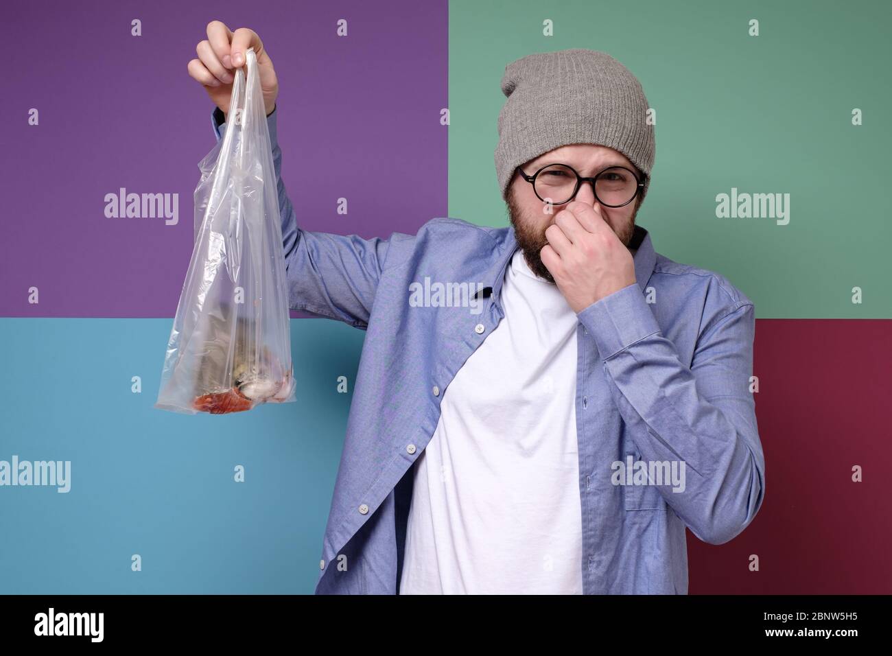 Caucasian man holds a stinky, spoiled fish in a transparent package and pinches nose with his fingers due to an unpleasant odor. Stock Photo