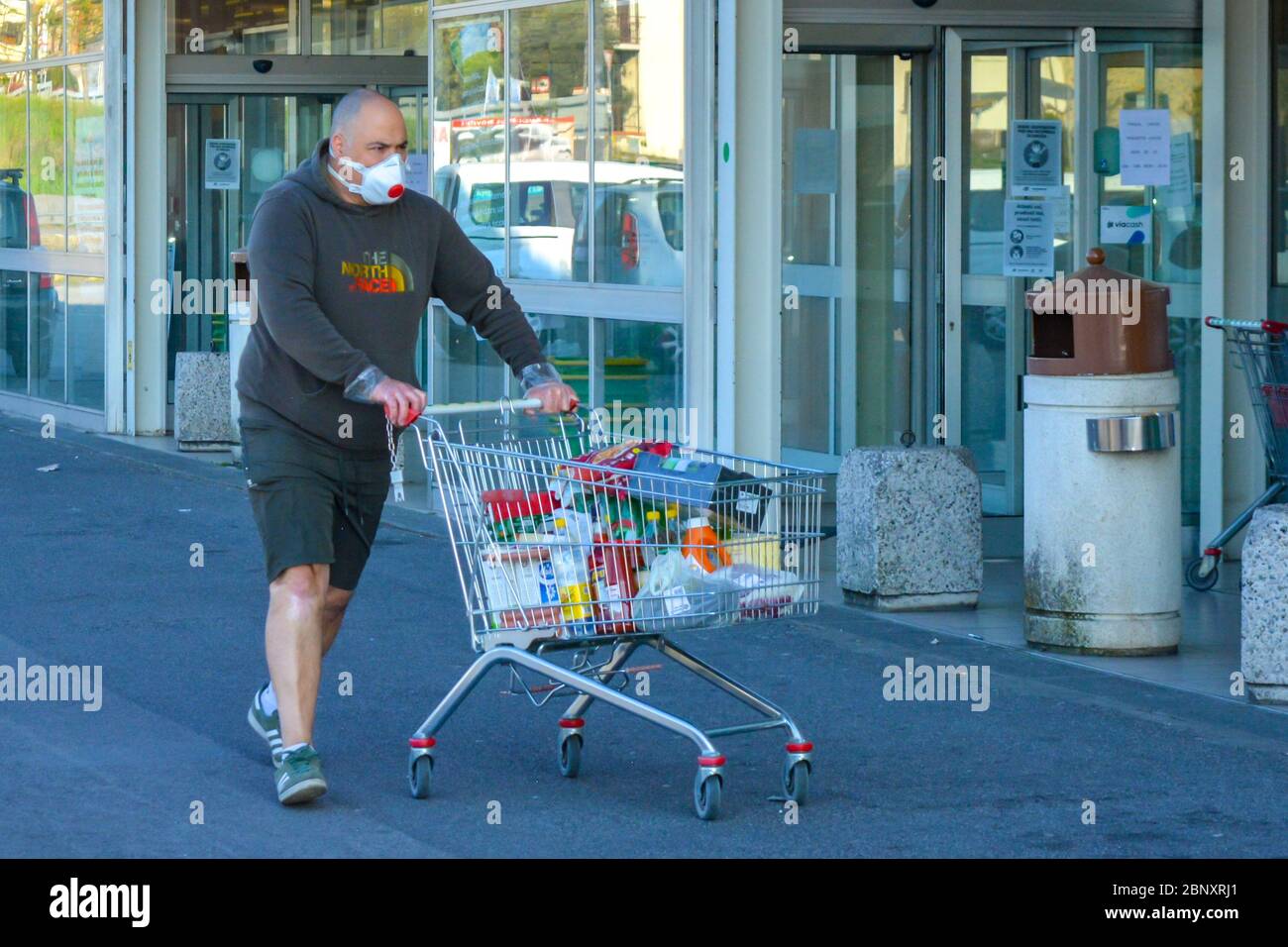 Closeup of a serious looking man with coronavirus protective mask pushing his shopping cart full of essentials. Routine scene outside grocery stores d Stock Photo