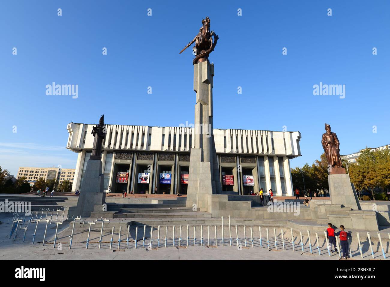 Brutalist style Kyrgyz National Philharmonic in Bishkek, Kyrgyzstan, named in honor of musician Toktogul Satylganov. Red flowers and Manas Statue. Stock Photo
