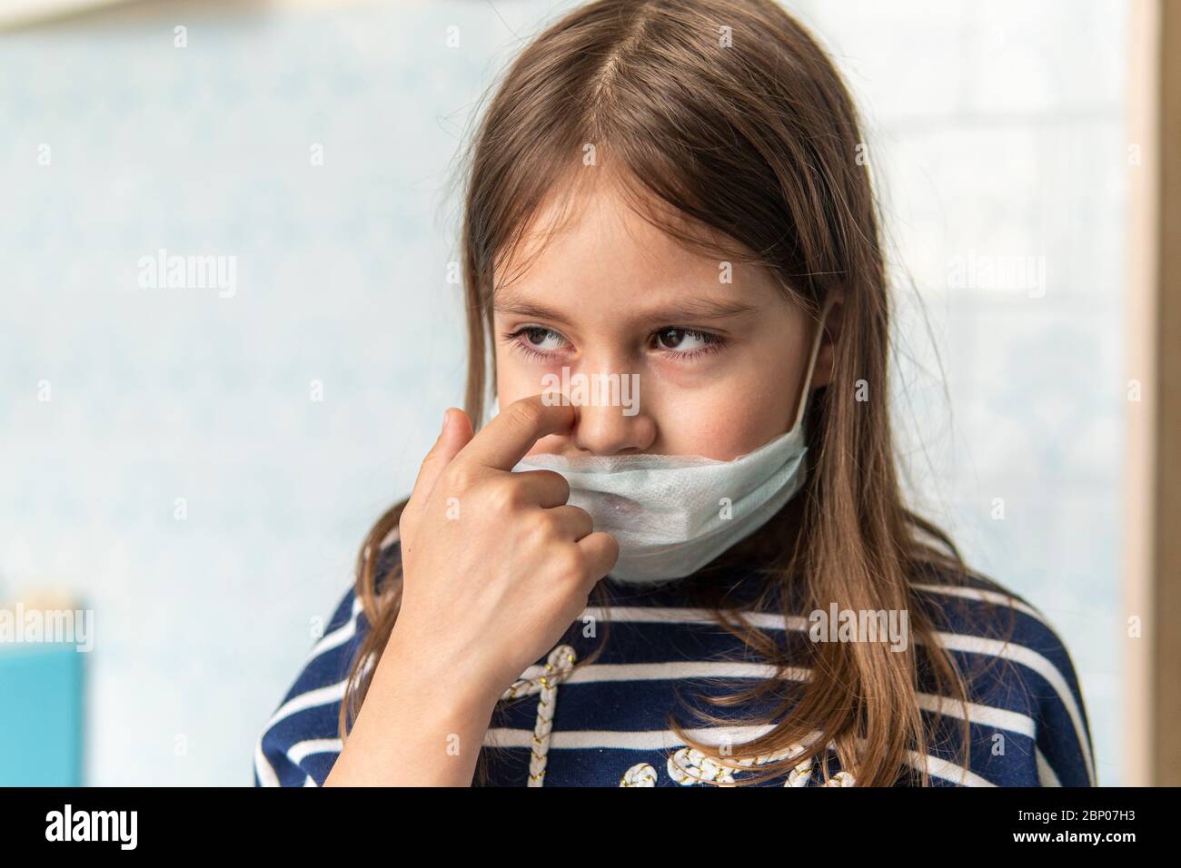 Toddler girl with medical mask on face. Covid19 corona virus outbreak. girl touches face dirty hands, it's not safe. Sad girl is tired of sitting at h Stock Photo