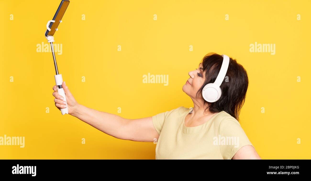 Beautiful Senior woman making video call by smartphone, monopod and wireless headphones over yellow background. Stock Photo