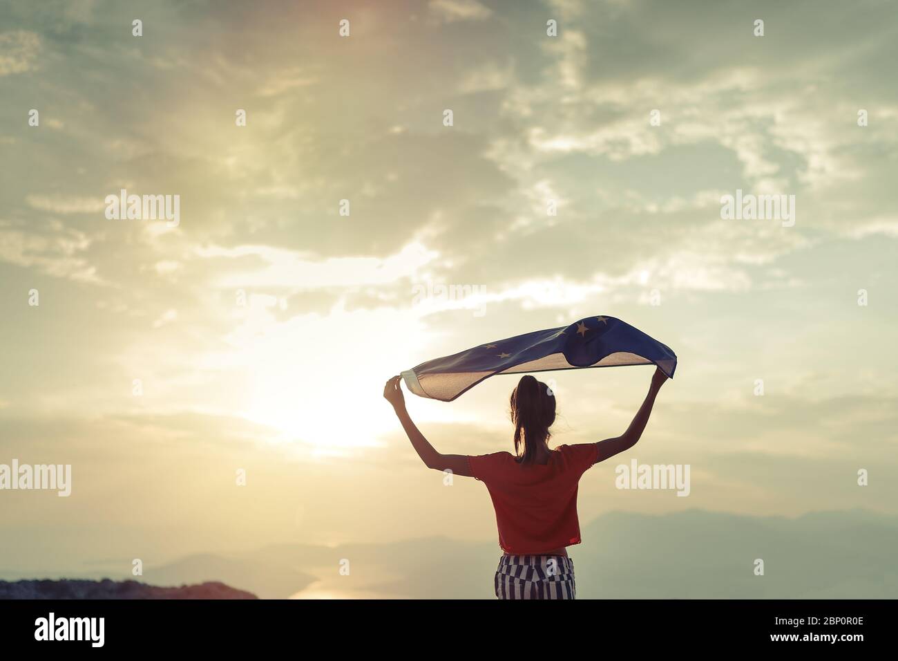 Child girl teenager young person is waving European Union flag on top of mountain at sky background. Sunset time Stock Photo