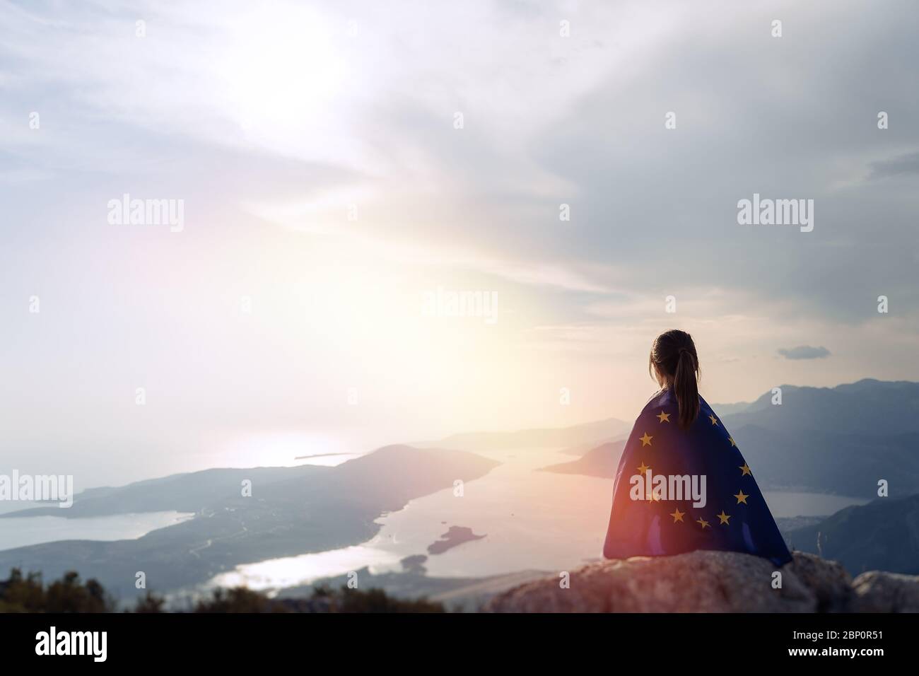 Child teenager girl sitting on the top of the mountain with an European Union flag on her shoulders. Sunset time Stock Photo